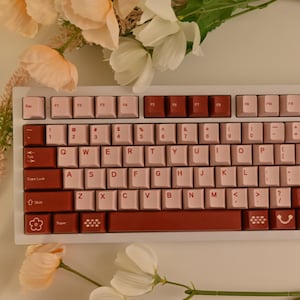 May include: A white mechanical keyboard with red and pink keycaps. The keyboard has a number row, function keys, and a row of keys with symbols. The keyboard is surrounded by pink flowers.