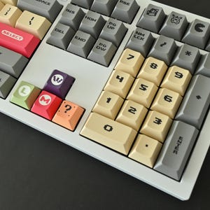 May include: A mechanical keyboard featuring a color-blocked layout with cream, gray, red, purple, green, and orange keycaps. The keys display pixelated fonts, numbers, and various icons including a star, a ghost, and a question mark.