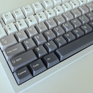 May include: A close-up of a mechanical keyboard with a gradient keycap design. The keys transition from white to gray to dark gray. The keyboard has a white frame and features standard alphanumeric keys, function keys, and modifier keys.
