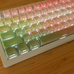 May include: A mechanical computer keyboard featuring a gradient color scheme that transitions from soft green on the left side to pale pink on the right side. The keys are backlit with white light, and the frame is a solid, neutral off-white color.