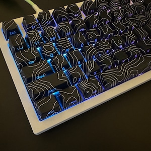 May include: A white mechanical keyboard with black keycaps featuring a topographic pattern. The keys are backlit with a blue light. The keyboard has keys labeled 'Esc', 'Tab', 'Caps', 'Shift', 'Ctrl', 'Win', 'Alt', 'F1', 'F2', 'F3', 'F4', 'F5', 'F6', 'F7', 'F8', 'F9', 'F10', 'F11', 'F12', 'Q', 'W', 'E', 'R', 'T', 'Y', 'U', 'I', 'O', 'P', 'A', 'S', 'D', 'F', 'G', 'H', 'J', 'K', 'L', 'Z', 'X', 'C', 'V', 'B', 'N', 'M', '1', '2', '3', '4', '5', '6', '7', '8', '9', '0', '-', '=', '[', ']', ';', '"', ',', '.', '/', 'Backspace', 'Enter', 'Space', 'Delete', 'Insert', 'Home', 'Page Up', 'End', 'Page Down', 'Arrow Up', 'Arrow Down', 'Arrow Left', 'Arrow Right'.
