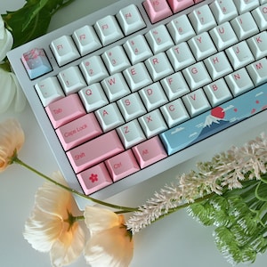 May include: A white mechanical keyboard with pink and blue keycaps. The keyboard has a Japanese-inspired design with a mountain and cherry blossom print on the space bar and a red sun on the enter key.