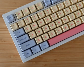 May include: A white mechanical keyboard with cream-colored keys and a pink space bar. The keys have red lettering and are labeled with the standard QWERTY layout.