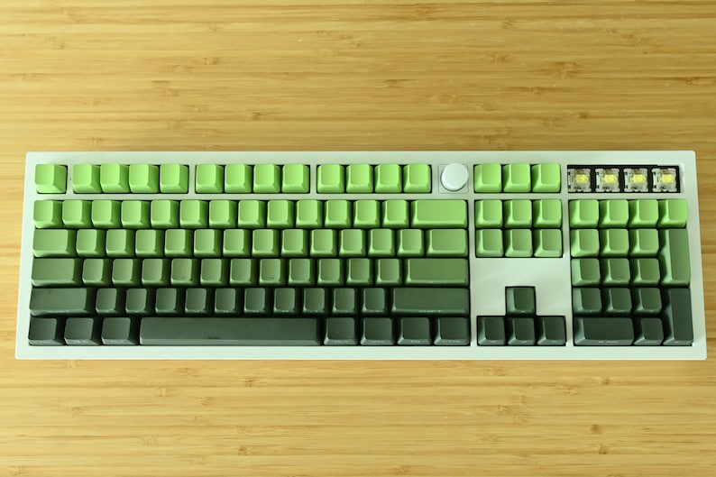 以下が含まれることがあります： 緑と黒のキーキャップが付いた白いメカニカルキーボード。キーキャップは、明るい緑から暗い緑へのグラデーションカラーになっています。キーボードには丸いノブと4つの追加キーの列があります。