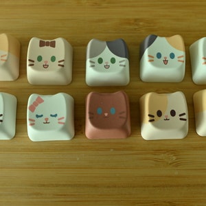 May include: Ten different keycaps featuring cute cartoon cats with various expressions and colours. The keycaps are made of white plastic with a glossy finish.