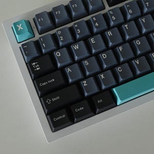 May include: A black and white mechanical keyboard with a teal space bar and escape key. The keyboard has a silver frame and black keys with white lettering. The keys are labelled with letters, numbers, and symbols.