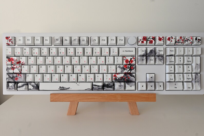May include: A white mechanical keyboard with a Japanese-inspired design. The keys have a black and white ink wash design with red cherry blossoms. The keyboard is on a wooden stand.