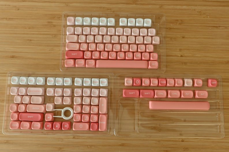 K&ouml;nnte beinhalten: Ein Satz rosa und wei&szlig;er Tastaturkappen in einem durchsichtigen Kunststoffgeh&auml;use. Die Tastenkappen sind in Reihen und Spalten angeordnet, mit den Buchstaben des Alphabets, Zahlen und Symbolen darauf gedruckt. Die Tastenkappen sind in einem Farbverlauf von Rosa, von Hellrosa bis Dunkelrosa.