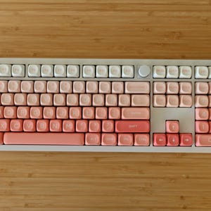 K&ouml;nnte beinhalten: Eine wei&szlig;e Tastatur mit rosa und wei&szlig;en Tastenkappen. Die Tastenkappen sind in einem Farbverlauf von rosa gehalten, wobei das hellste Rosa oben und das dunkelste Rosa unten ist. Die Tastatur hat einen wei&szlig;en Rahmen und eine wei&szlig;e Basis.