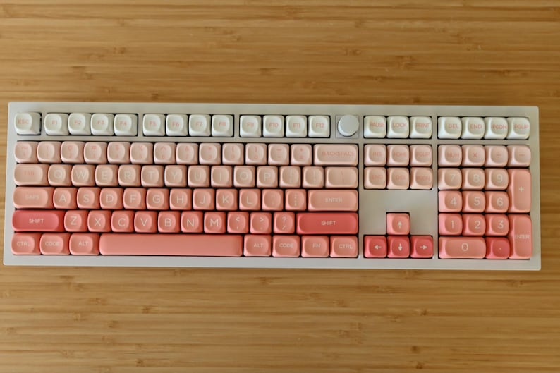 K&ouml;nnte beinhalten: Eine wei&szlig;e Tastatur mit rosa und wei&szlig;en Tastenkappen. Die Tastenkappen sind in einem Farbverlauf von rosa gehalten, wobei das hellste Rosa oben und das dunkelste Rosa unten ist. Die Tastatur hat einen wei&szlig;en Rahmen und eine wei&szlig;e Basis.