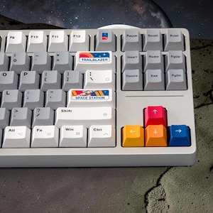 May include: A light gray mechanical keyboard featuring white and gray keycaps. Specific keys display space-themed graphics and the text TRAILBLAZER and SPACE STATION. The arrow keys are colored in bright pink, yellow, orange, and blue.