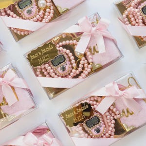 May include: A clear gift box containing a pink digital tally counter, a string of pink pearls, and a small pink velvet pouch. A gold-colored plaque on the box features the text Aman & Amanda 25.12.25. The box is tied with a light pink satin ribbon.