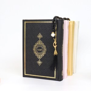 May include: A black book with gold ornate detailing and a matching gold border. A black beaded prayer beads with a gold charm and tassel hangs over the book. Other books in pink, gold, and white are visible.