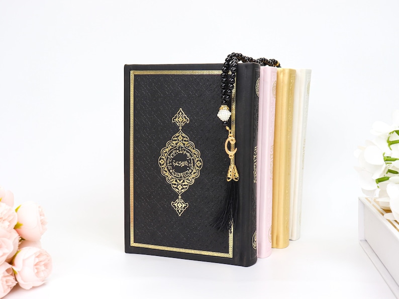 May include: A black book with gold ornate detailing and a matching gold border. A black beaded prayer beads with a gold charm and tassel hangs over the book. Other books in pink, gold, and white are visible.