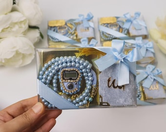 May include: A clear box contains a light blue beaded prayer beads set with a digital counter, a small blue book, and a gold tassel. The box is tied with a light blue ribbon. Several similar boxes are visible in the background.