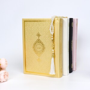 May include: A stack of religious books in gold, white, black, and pink. The gold book has an ornate design and Arabic text. A pearl rosary with a white tassel hangs over the books. Artificial flowers are in the foreground.