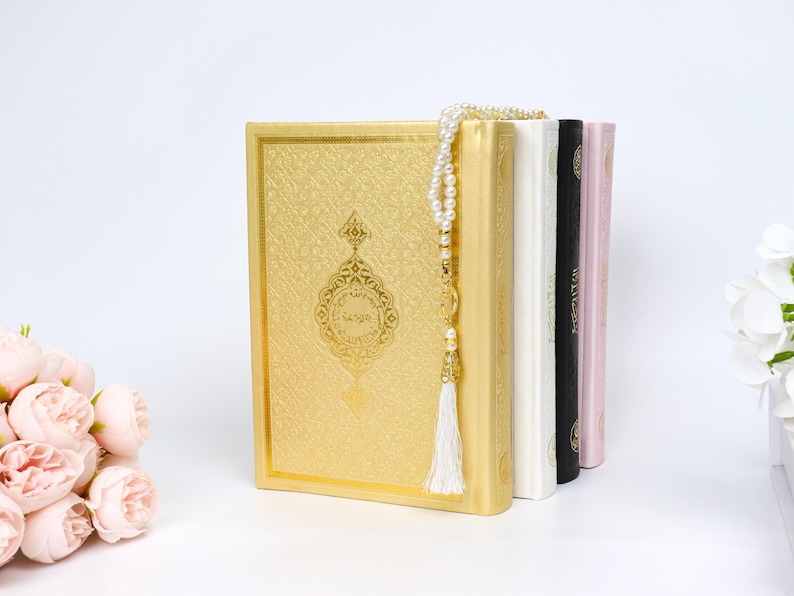 May include: A stack of religious books in gold, white, black, and pink. The gold book has an ornate design and Arabic text. A pearl rosary with a white tassel hangs over the books. Artificial flowers are in the foreground.