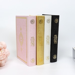May include: Four hardcover books in pink, gold, white, and black, standing upright. The books have gold embossed designs and Arabic script. A bouquet of pink flowers is in the lower left corner.