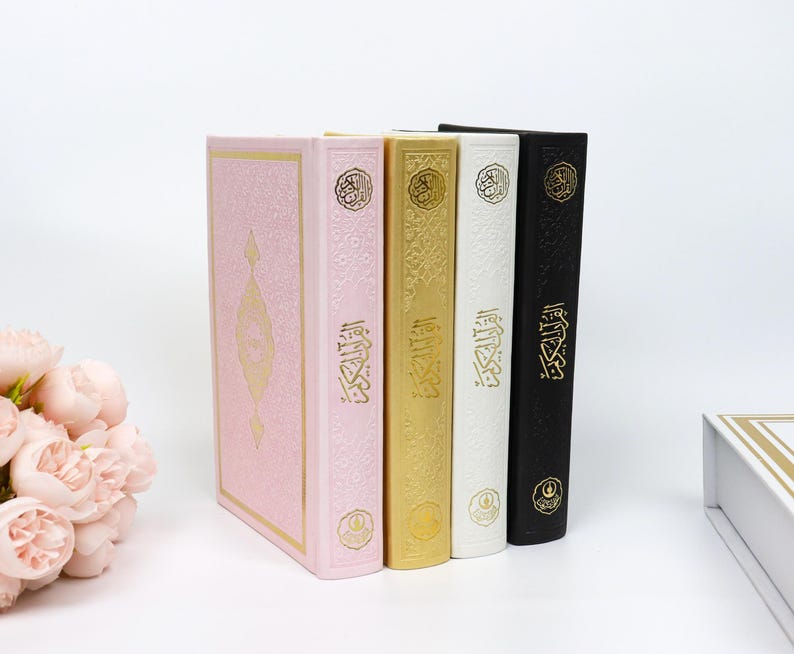 May include: Four hardcover books in pink, gold, white, and black, standing upright. The books have gold embossed designs and Arabic script. A bouquet of pink flowers is in the lower left corner.
