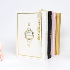 May include: A stack of religious books in various colors, including white, black, pink, and gold. The white book features gold ornate detailing and a gold-colored tassel. A string of pearls hangs over the books. Pink flowers are in the background.