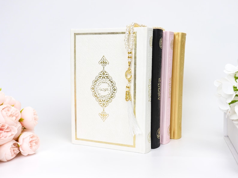 May include: A stack of religious books in various colors, including white, black, pink, and gold. The white book features gold ornate detailing and a gold-colored tassel. A string of pearls hangs over the books. Pink flowers are in the background.