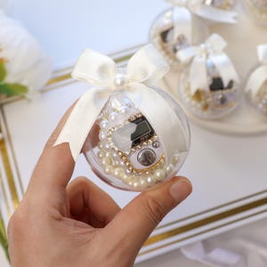 Op de afbeelding: Een doorzichtige plastic bal gevuld met parels en een digitale teller, vastgebonden met een wit lint en een parel. Er staan ​​verschillende soortgelijke ballen op de achtergrond. De bal wordt in een hand gehouden.