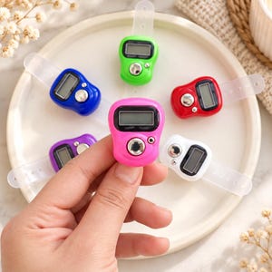 Puede incluir: Una colección de contadores digitales de dedo con una pequeña pantalla LCD y un botón plateado, disponibles en colores brillantes como rosa, azul, verde, rojo y blanco, cada uno equipado con una correa de silicona transparente ajustable.
