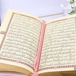 May include: An open book with Arabic script, likely the Quran, featuring ornate red and gold borders. A string of pink beads serves as a bookmark. The pages are cream-colored, and the book is set against a soft background.