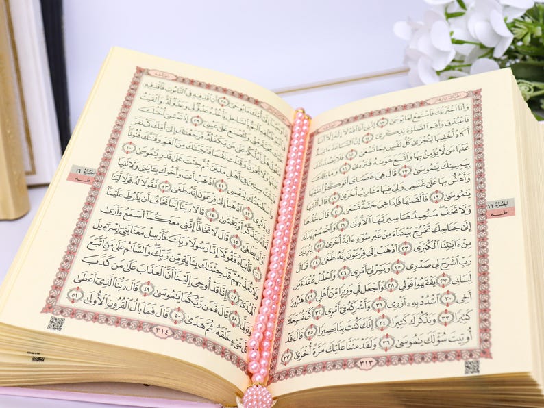 May include: An open book with Arabic script, likely the Quran, featuring ornate red and gold borders. A string of pink beads serves as a bookmark. The pages are cream-colored, and the book is set against a soft background.
