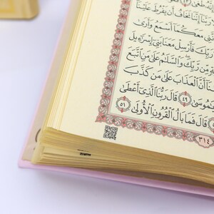 May include: Open book with Arabic script. The pages are cream-colored with black text and a decorative red and gold border. The book has a pink cover and a QR code is visible on one page.