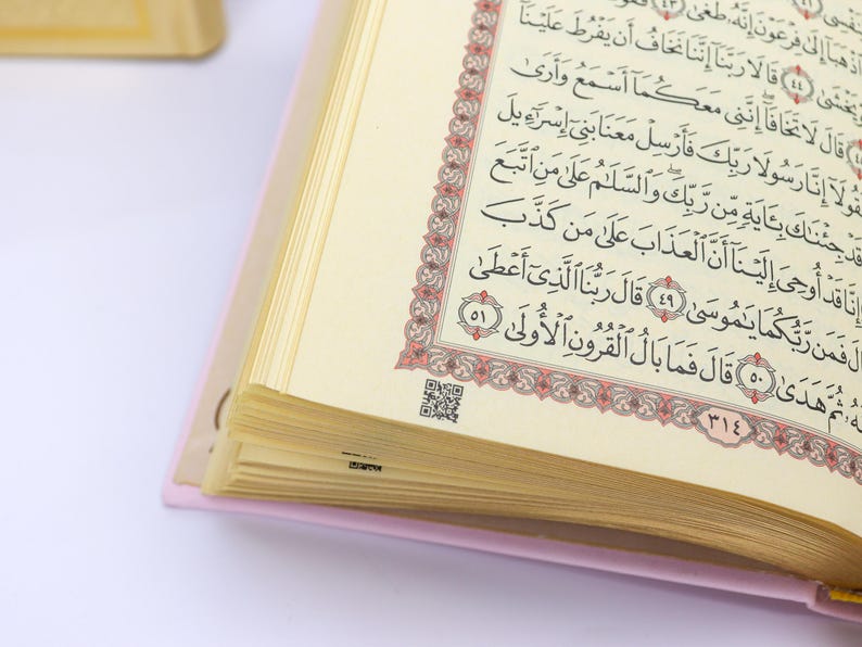 May include: Open book with Arabic script. The pages are cream-colored with black text and a decorative red and gold border. The book has a pink cover and a QR code is visible on one page.