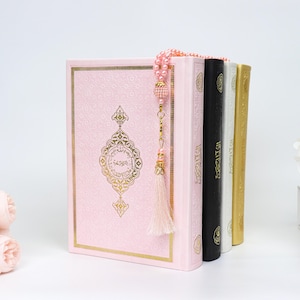 May include: A stack of religious books, including a pink book with gold accents and a decorative design. A beaded strand with a tassel hangs over the books. Additional books are black, white, and gold. A bouquet of pink flowers is in the foreground.