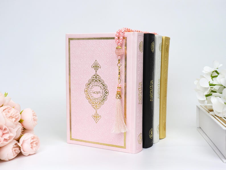 May include: A stack of religious books, including a pink book with gold accents and a decorative design. A beaded strand with a tassel hangs over the books. Additional books are black, white, and gold. A bouquet of pink flowers is in the foreground.