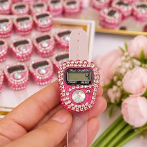 Könnte beinhalten: Ein leuchtend pinkfarbener digitaler Tally-Zähler mit einem verstellbaren, transparenten Kunststoffarmband. Das Gerät verfügt über ein kleines LCD-Display mit der Zahl 4, einen großen schillernden runden Knopf und eine Reset-Taste, umrandet von Strass.