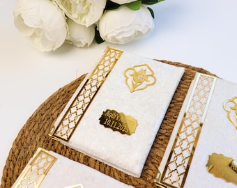 May include: White fabric-covered books with gold accents and decorative gold embellishments. The books have a gold plaque with the text "Aqib's Aameen 10.11.2025". The books are arranged on a woven mat with white flowers in the background.