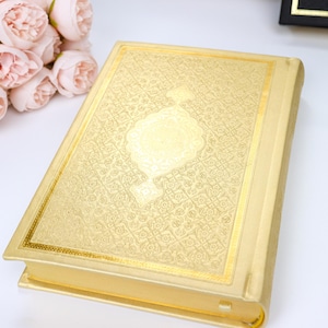 May include: A gold-colored book with an ornate, embossed design on the cover. The book has a rectangular shape with gold trim and a decorative central emblem. A bouquet of pink flowers is visible in the background.
