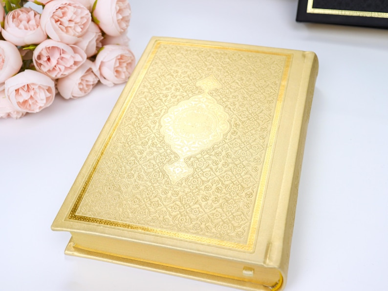 May include: A gold-colored book with an ornate, embossed design on the cover. The book has a rectangular shape with gold trim and a decorative central emblem. A bouquet of pink flowers is visible in the background.