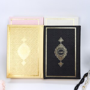 May include: Four books with gold and black covers are displayed. The gold cover has an ornate design. The black cover has gold text and a decorative emblem. Two smaller books in pink and white are behind the larger books.