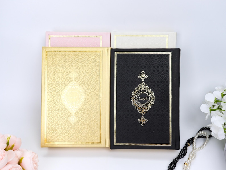 May include: Four books with gold and black covers are displayed. The gold cover has an ornate design. The black cover has gold text and a decorative emblem. Two smaller books in pink and white are behind the larger books.