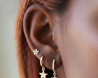 May include: Close-up of an ear with three gold earrings. One is a small flower stud, and the other two are hoop earrings with star pendants. One star pendant is plain gold, and the other is encrusted with small, clear stones.