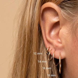 Puede incluir: Primer plano de una oreja con cuatro pendientes de aro dorados de diferentes tamaños: 12 mm, 14 mm, 16 mm y 18 mm. Los pendientes se llevan en la oreja, mostrando sus diferentes diámetros. El fondo es suave y neutro.
