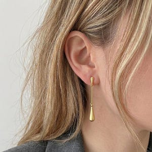 Peut inclure: Gros plan d'une boucle d'oreille dorée. La boucle d'oreille a un petit sommet arrondi qui s'attache à l'oreille, avec une longue barre fine qui mène à un pendentif en forme de goutte. La boucle d'oreille est suspendue à l'oreille d'une personne.