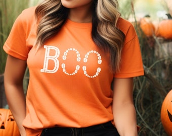 May include: A woman wearing an orange t-shirt with the word "BOO" spelled out in white with small circles. The circles are arranged to look like teeth. The woman is standing in front of a pile of pumpkins.