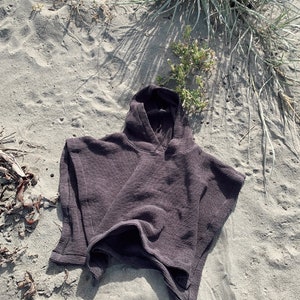 Puede incluir: Un poncho con capucha de color marr&oacute;n oscuro que presenta un tejido texturizado de punto de gofre. La prenda tiene capucha, mangas cortas y anchas, y una silueta holgada y cuadrada con un dobladillo recto, mostrando un patr&oacute;n de tejido en rejilla.