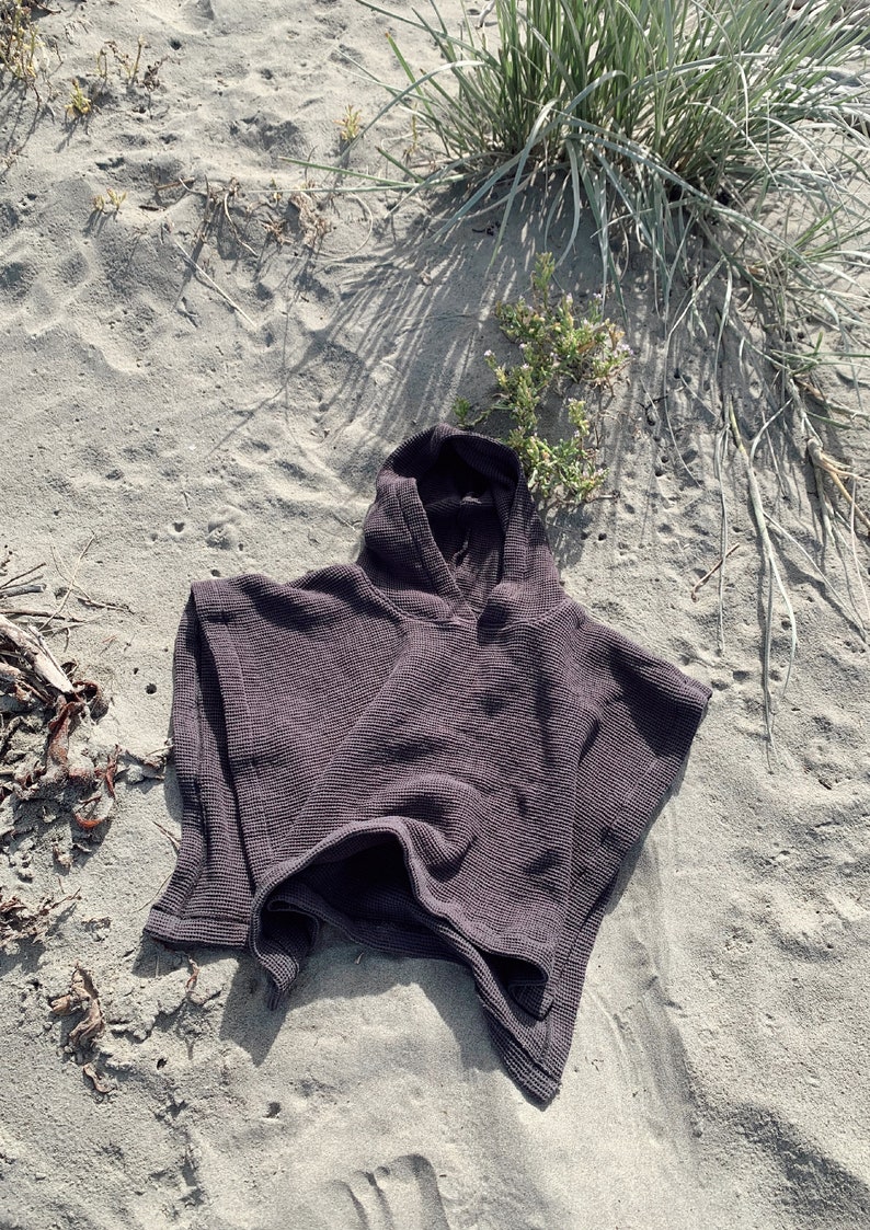 Puede incluir: Un poncho con capucha de color marr&oacute;n oscuro que presenta un tejido texturizado de punto de gofre. La prenda tiene capucha, mangas cortas y anchas, y una silueta holgada y cuadrada con un dobladillo recto, mostrando un patr&oacute;n de tejido en rejilla.