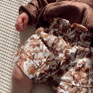 May include: A pair of brown and white floral print shorts with a frilled waistband. The shorts are made from a soft, lightweight fabric and have a relaxed fit.