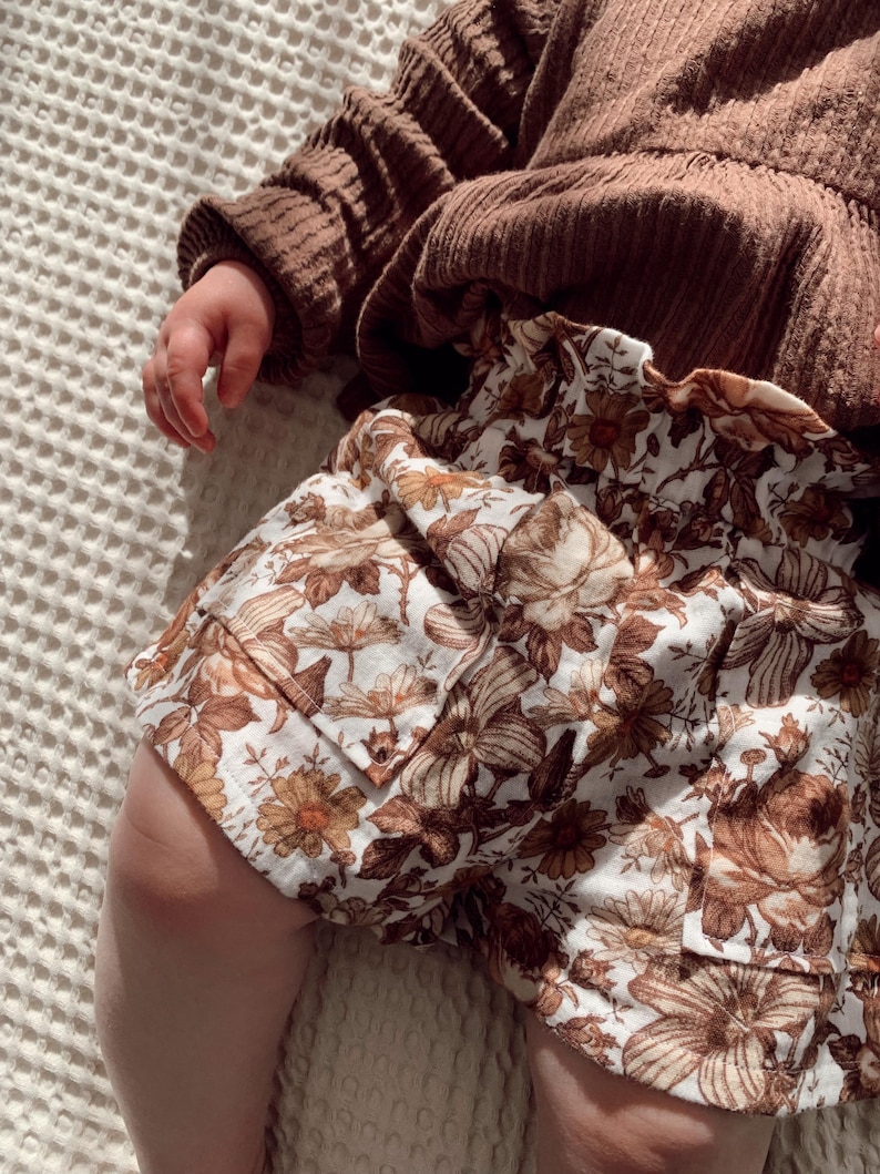 May include: A pair of brown and white floral print shorts with a frilled waistband. The shorts are made from a soft, lightweight fabric and have a relaxed fit.