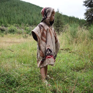 Puede incluir: Un poncho con capucha que presenta un patr&oacute;n geom&eacute;trico en crema, marr&oacute;n y negro con detalles en rojo. La prenda tiene una silueta holgada sin mangas con capucha incorporada, mostrando una textura tejida y un dise&ntilde;o de motivo repetitivo.