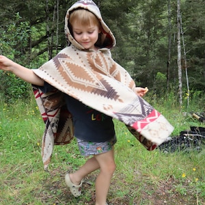 Puede incluir: Un poncho con capucha que presenta un patr&oacute;n geom&eacute;trico en beige, marr&oacute;n y rojo. La prenda se lleva sobre una camiseta oscura y pantalones cortos estampados, combinada con zuecos claros. El poncho tiene un corte amplio y drapeado con capucha.