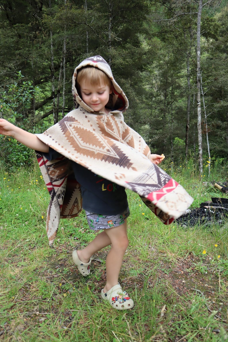 Puede incluir: Un poncho con capucha que presenta un patr&oacute;n geom&eacute;trico en beige, marr&oacute;n y rojo. La prenda se lleva sobre una camiseta oscura y pantalones cortos estampados, combinada con zuecos claros. El poncho tiene un corte amplio y drapeado con capucha.